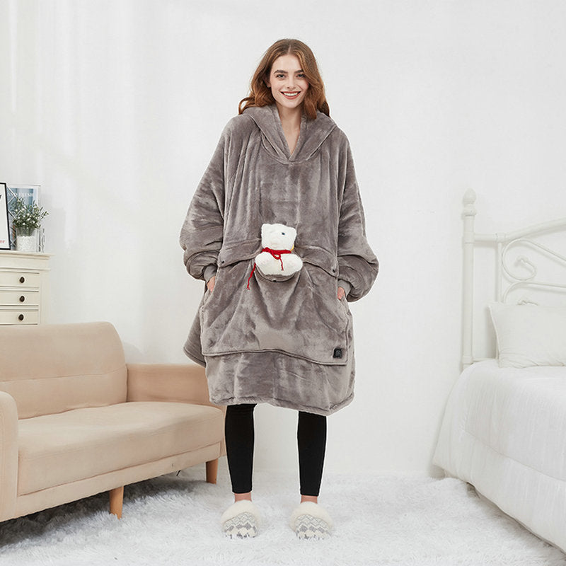 Woman wearing gray CozyGlow wearable heated hoodie blanket with front pocket, standing in a cozy bedroom